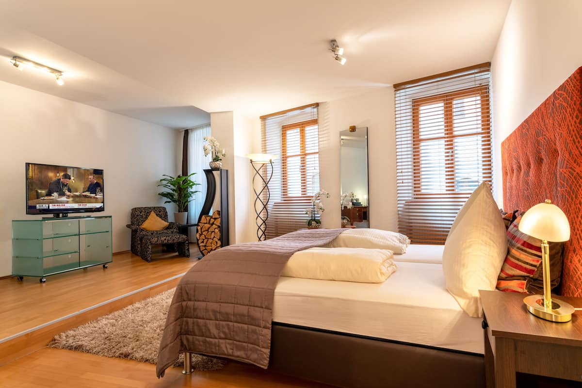 A modern bedroom with a bed, a TV, a desk, and a window with blinds.