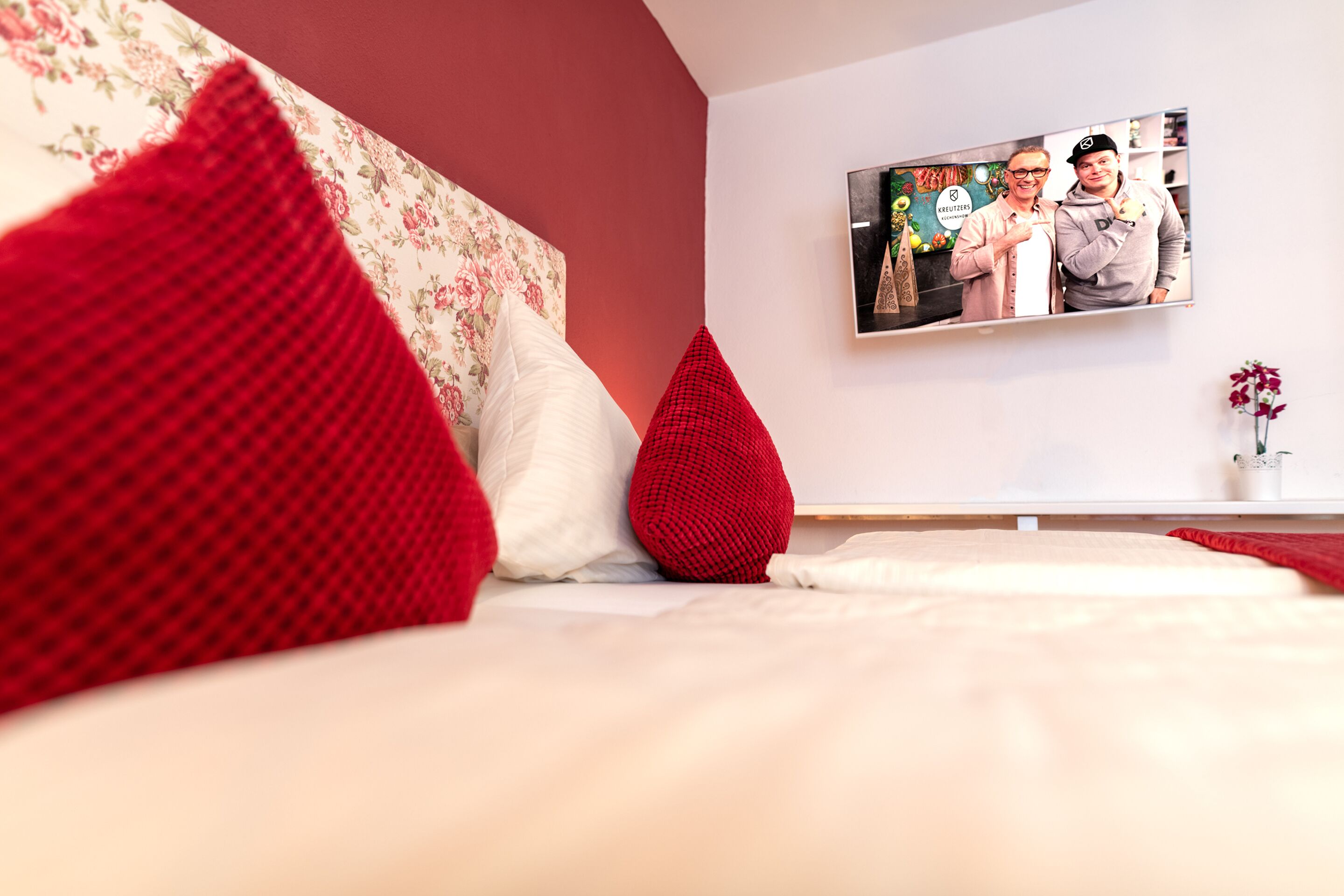 Een slaapkamer met een bed, rode kussens, een hoofdbord met bloemenprint, een flatscreen televisie aan de muur en een kleine vaas met bloemen.