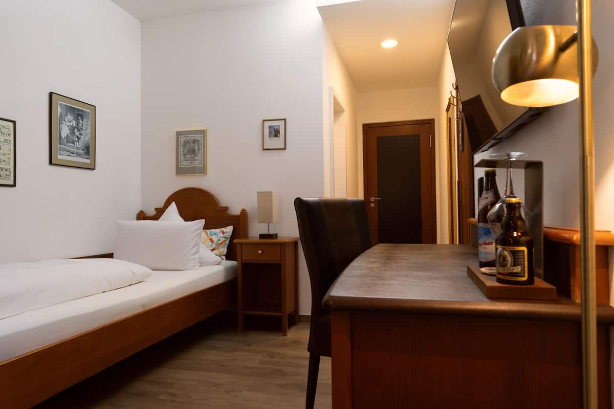 A hotel room with a bed, a desk, a chair, a lamp, and a bottle of beer on the desk.