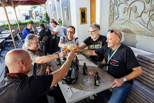 A group of people toasting with beer glasses at an outdoor wooden table in a restaurant setting.