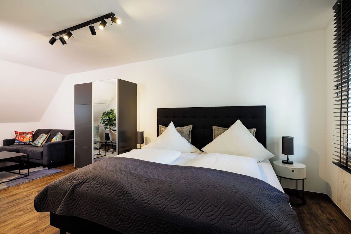 A modern bedroom with a large bed, a dark headboard, a bedside table with a lamp, and a view of a cityscape through the window.