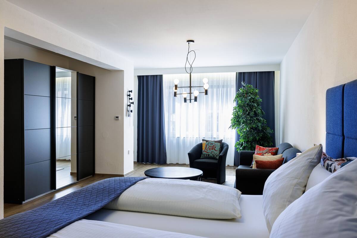 A modern hotel room with a large bed, a round coffee table, a sofa, a chair, and a large window with curtains.