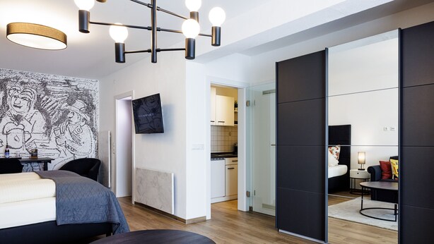 A modern hotel room with a bed, a round table, a TV, and a kitchen in the background. - Brauereigasthof Flair-Hotel Sperber-Bräu (Sulzbach-Rosenberg)