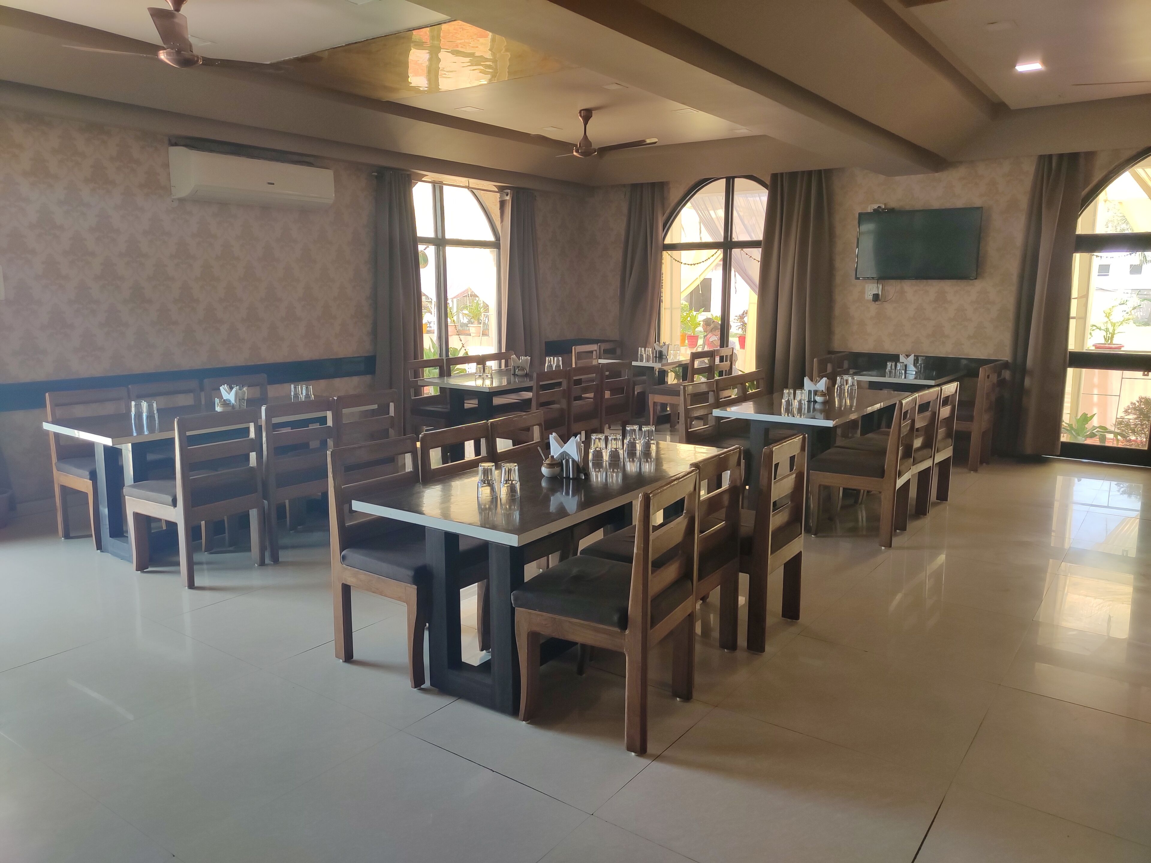 A spacious dining area with multiple wooden tables and chairs, large windows with curtains, and a flat-screen TV mounted on the wall.