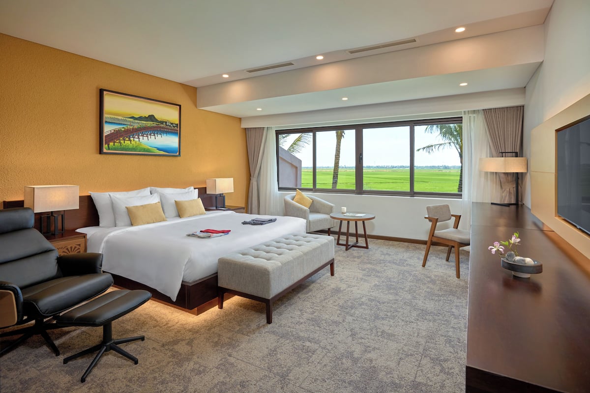 Ein Hotelzimmer mit einem großen Bett, einem Schreibtisch, einem Sessel und Blick auf ein grünes Feld.