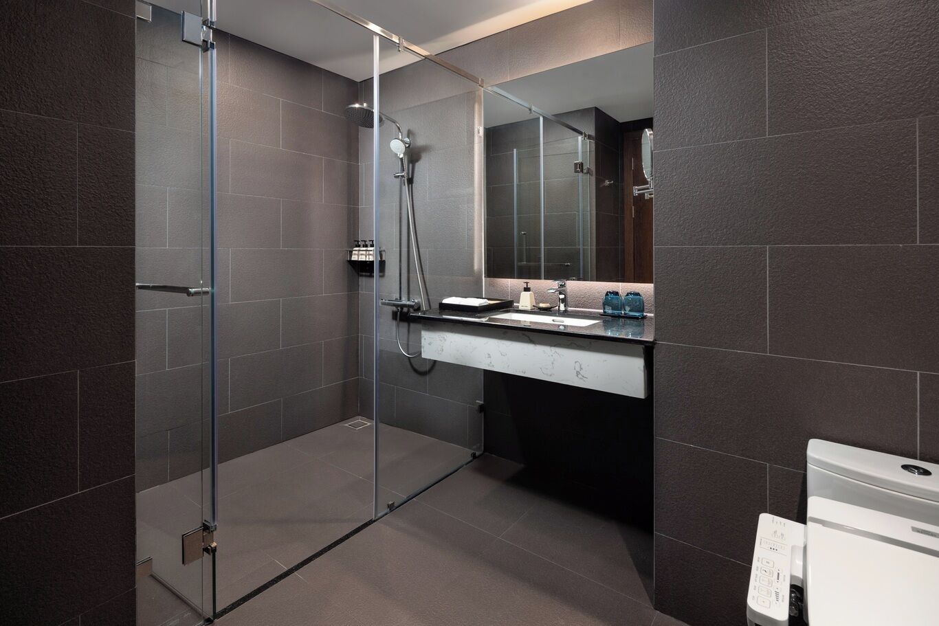 Et moderne badeværelse med et glasafskærmet brusebad, en vask med et spejl over, og et toilet med et kontrolpanel.