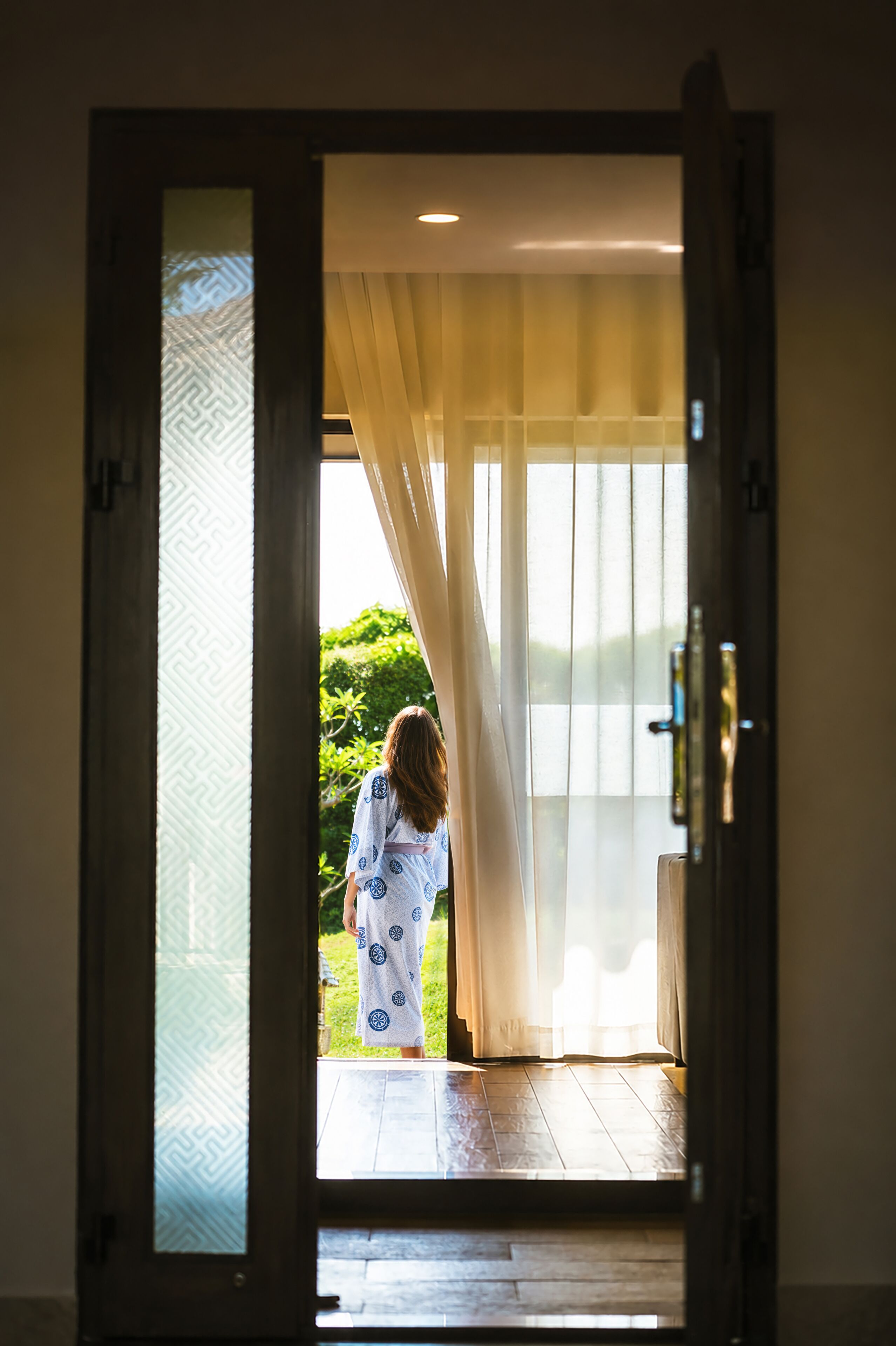 Un umbral con un panel de vidrio ornamental, una persona con un vestido estampado y vistas al exterior con vegetación.