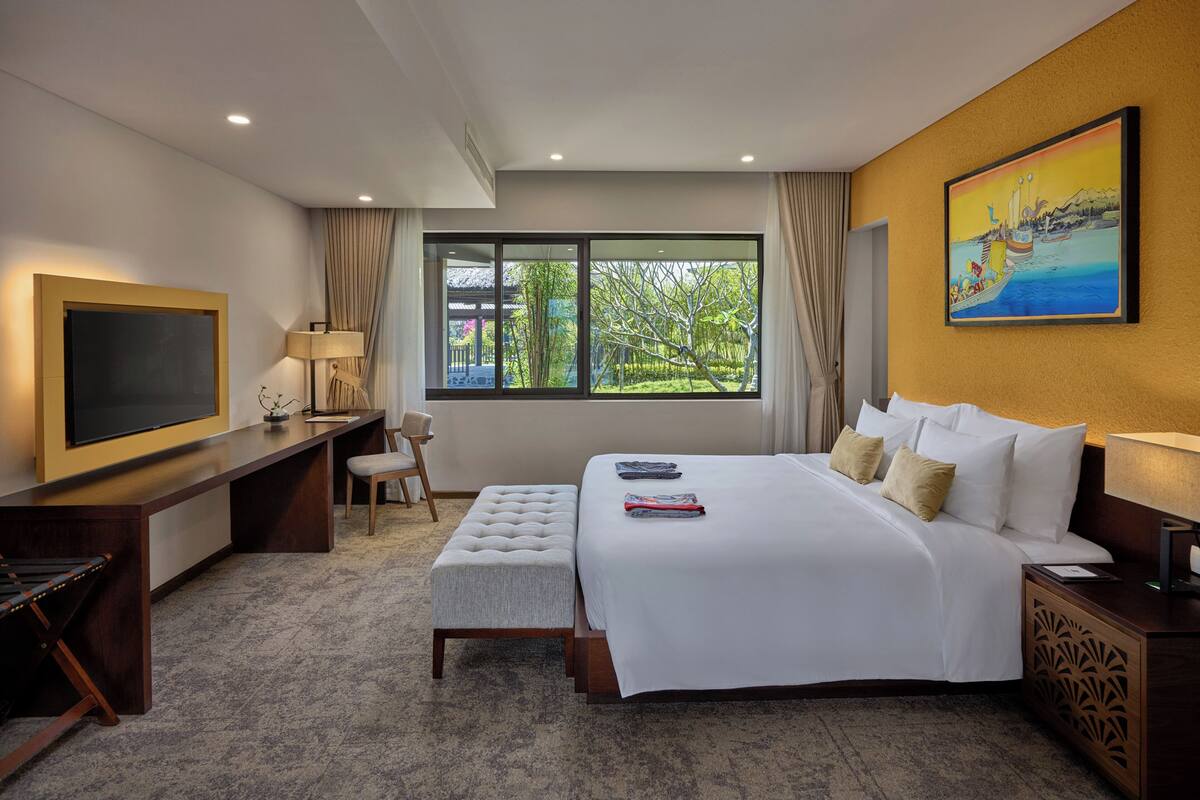 Ein Hotelzimmer mit einem großen Bett, einem Schreibtisch mit Fernseher, einem Sessel und einem Fenster mit Blick auf Grünflächen.