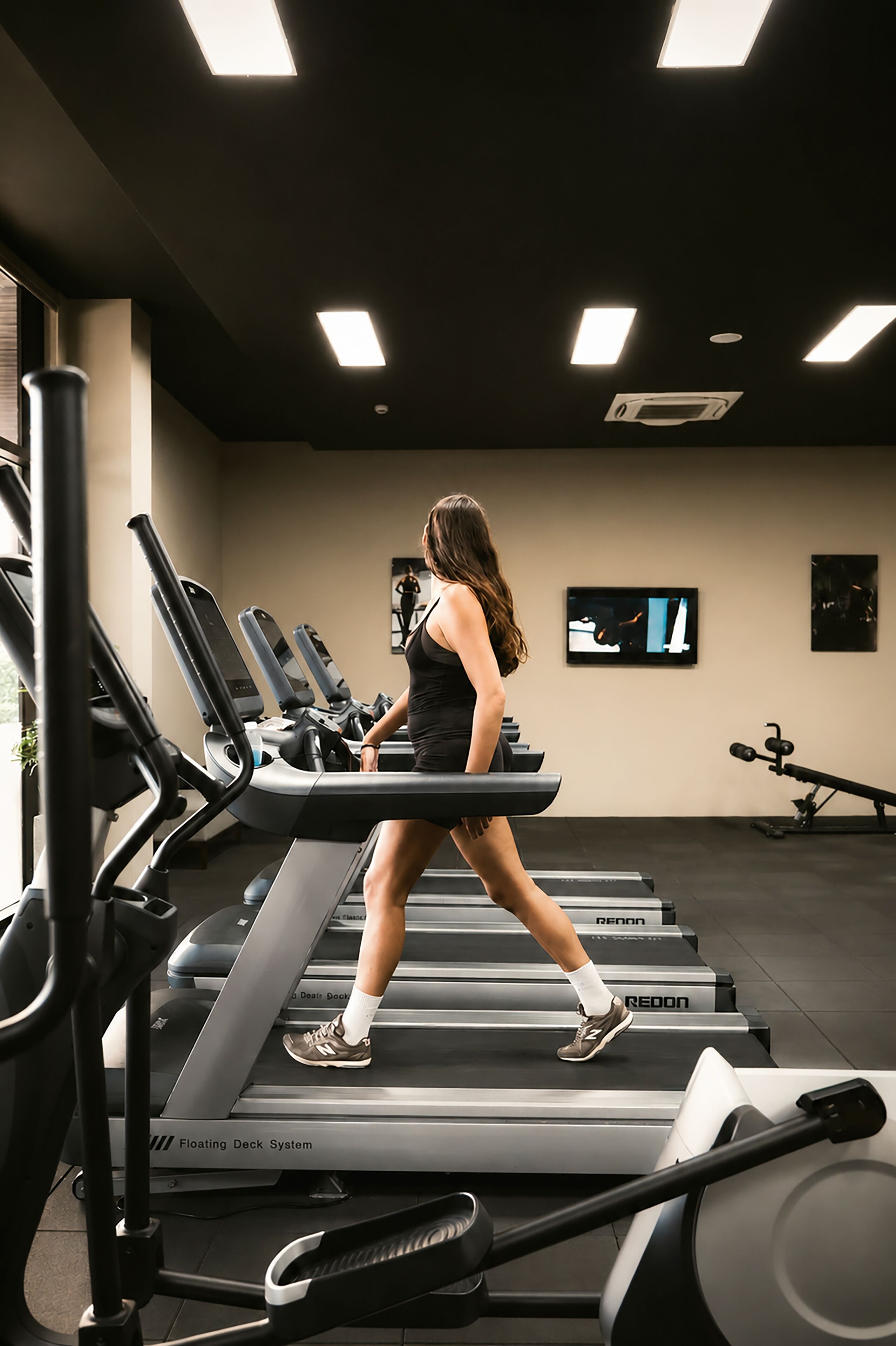 Una mujer caminando en una cinta de correr en un gimnasio con varias cintas, un televisor en la pared y equipo de ejercicio.