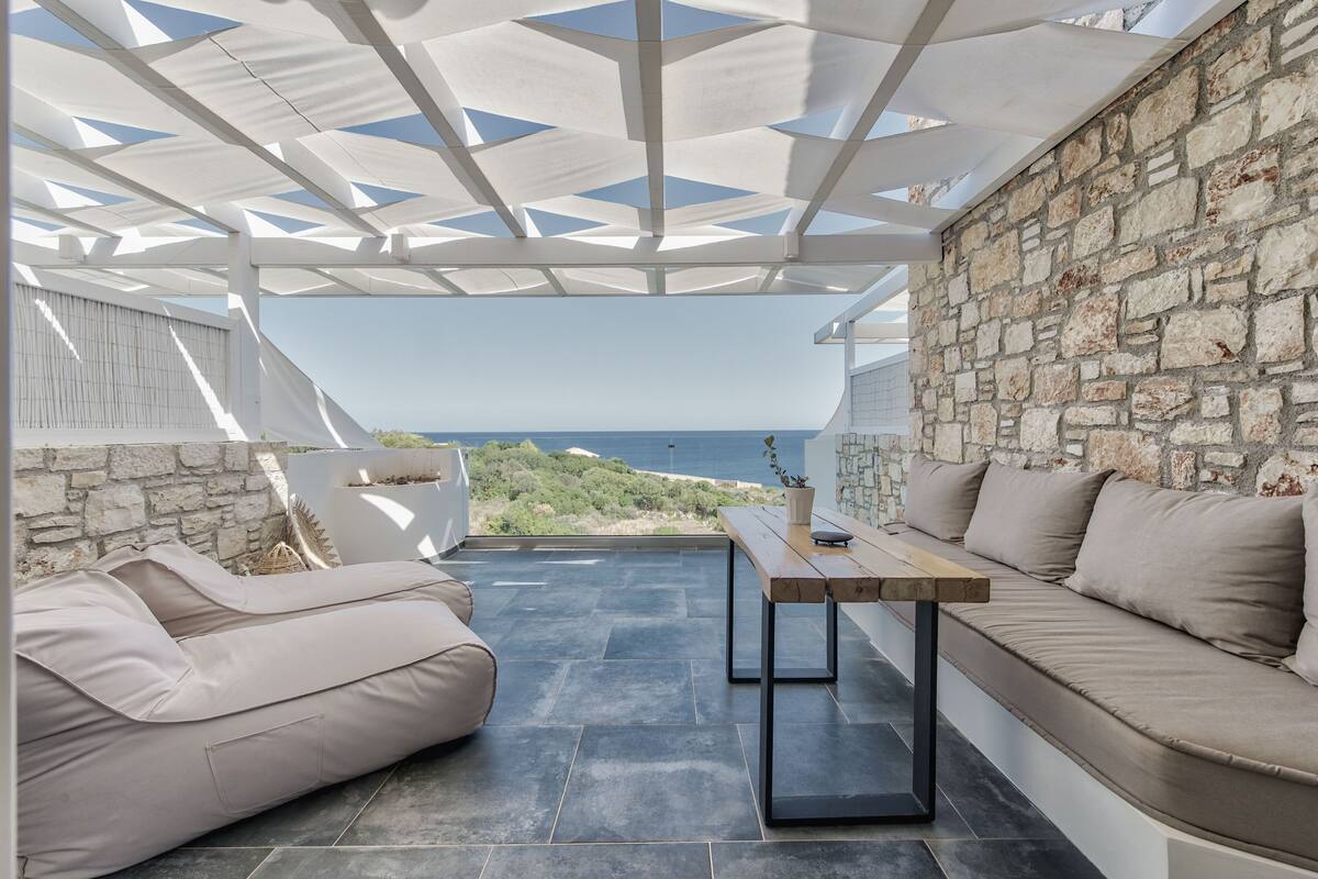 A covered outdoor seating area with stone walls, a view of the sea, and a wooden table.