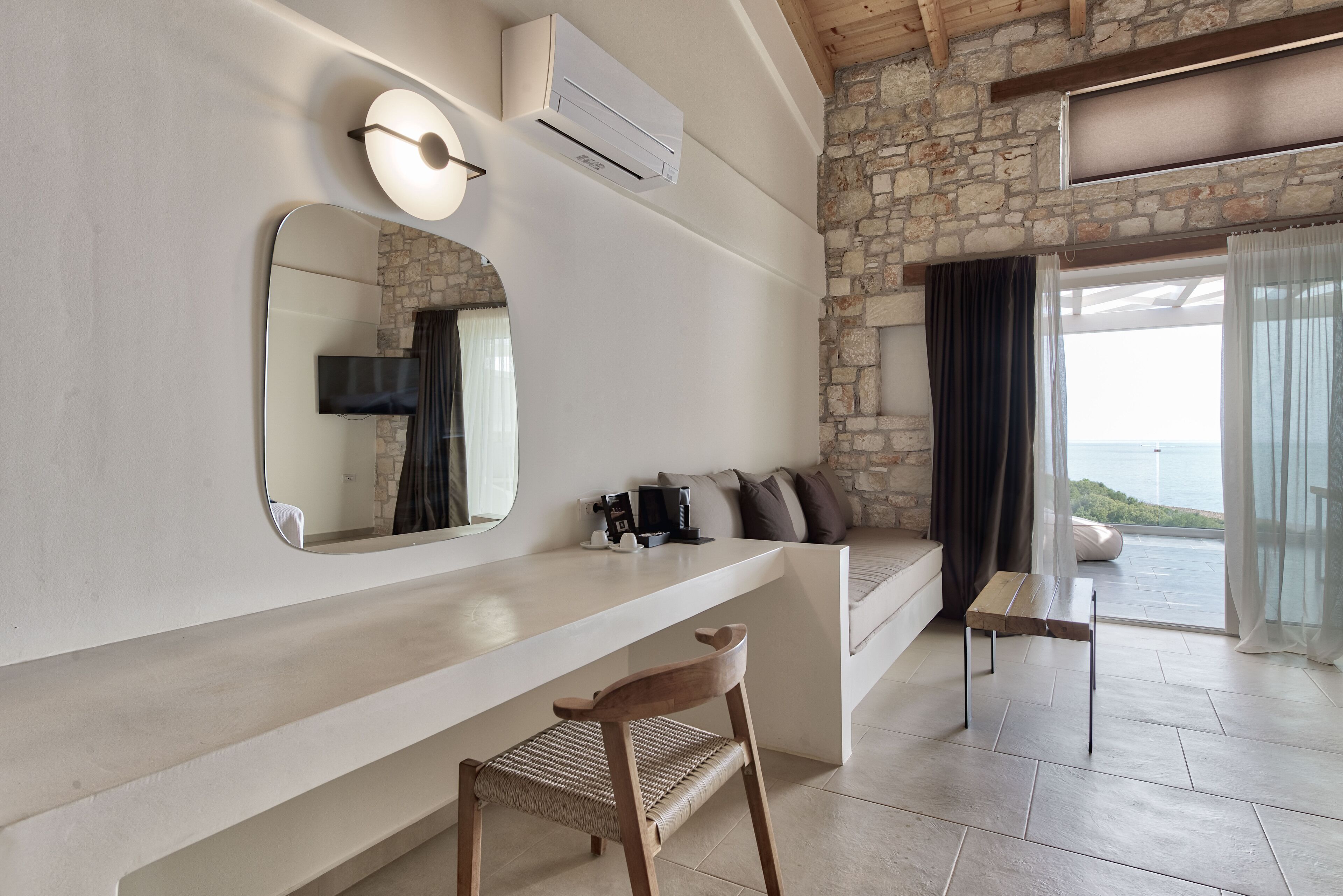 Una habitación moderna con pared de piedra, un espejo grande, televisor de pantalla plana, banca de madera y vista al mar.