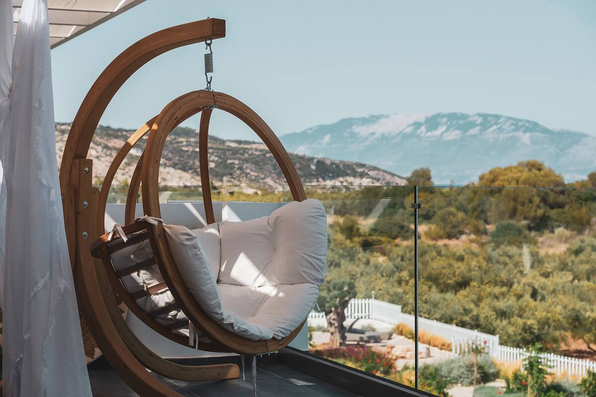 A wooden swing chair with a cushion, a glass railing, and a view of mountains and greenery.