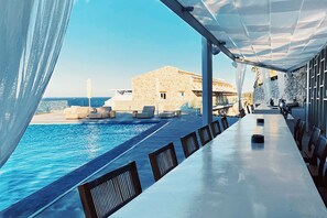 A poolside area with a long table and chairs, a view of the sea, and a building in the background. - Aliv Stone Suites & Spa (Zakynthos)