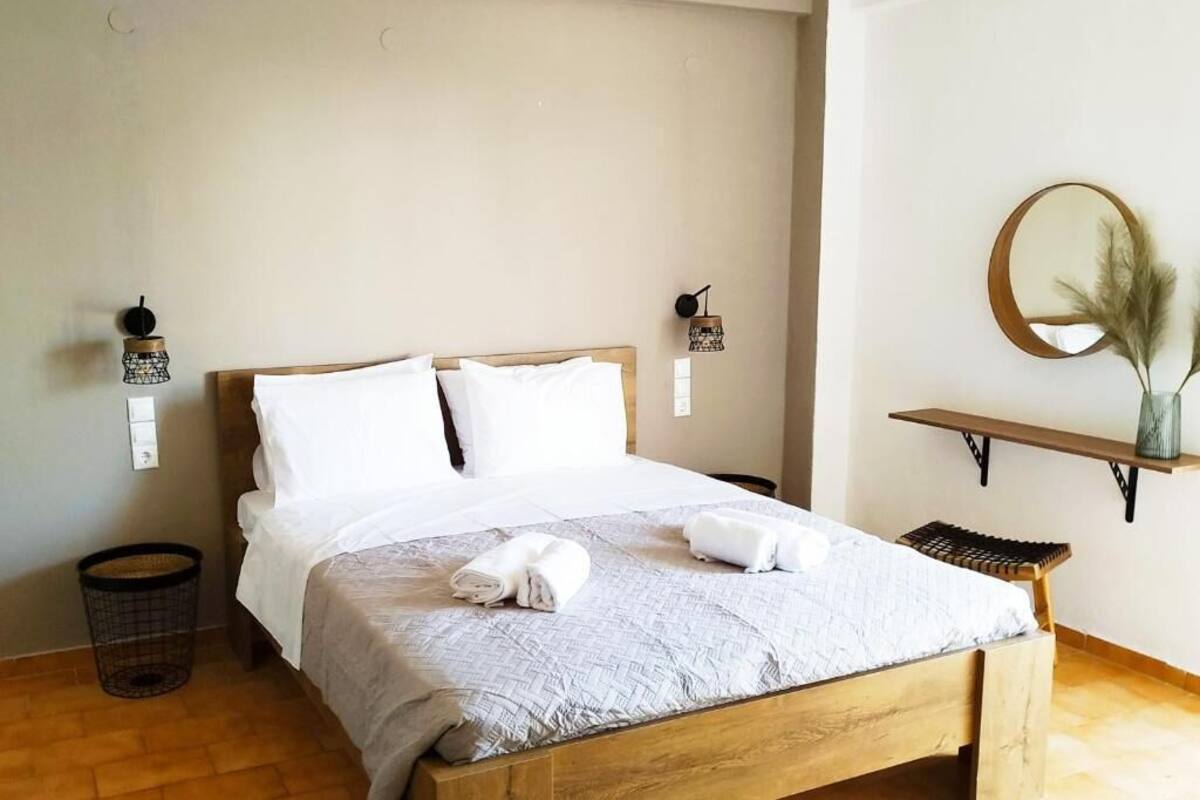 Cama bem arrumada com lençóis brancos e toalhas enroladas, cabeceira de madeira, um pequeno espelho redondo sobre uma prateleira e um banquinho de vime.