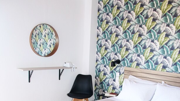 A neatly made bed with white linens and towels, a wooden headboard, a wall with a tropical leaf pattern, a round mirror, and a black chair. - Stalis Drops (Hersonissos)