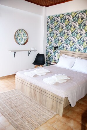 A neatly made bed with white linens and towels, a wooden headboard, a wall with a tropical leaf pattern, a round mirror, and a black chair. - Stalis Drops (Hersonissos)