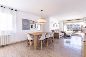 Un moderno salón con una mesa de comedor de madera, sillas grises, un sofá y un ventanal con vistas a edificios.