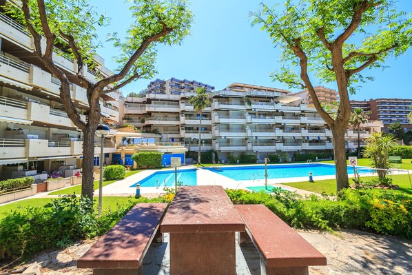 Zona de piscina con un banco, rodeada de vegetación y árboles, en un complejo residencial.