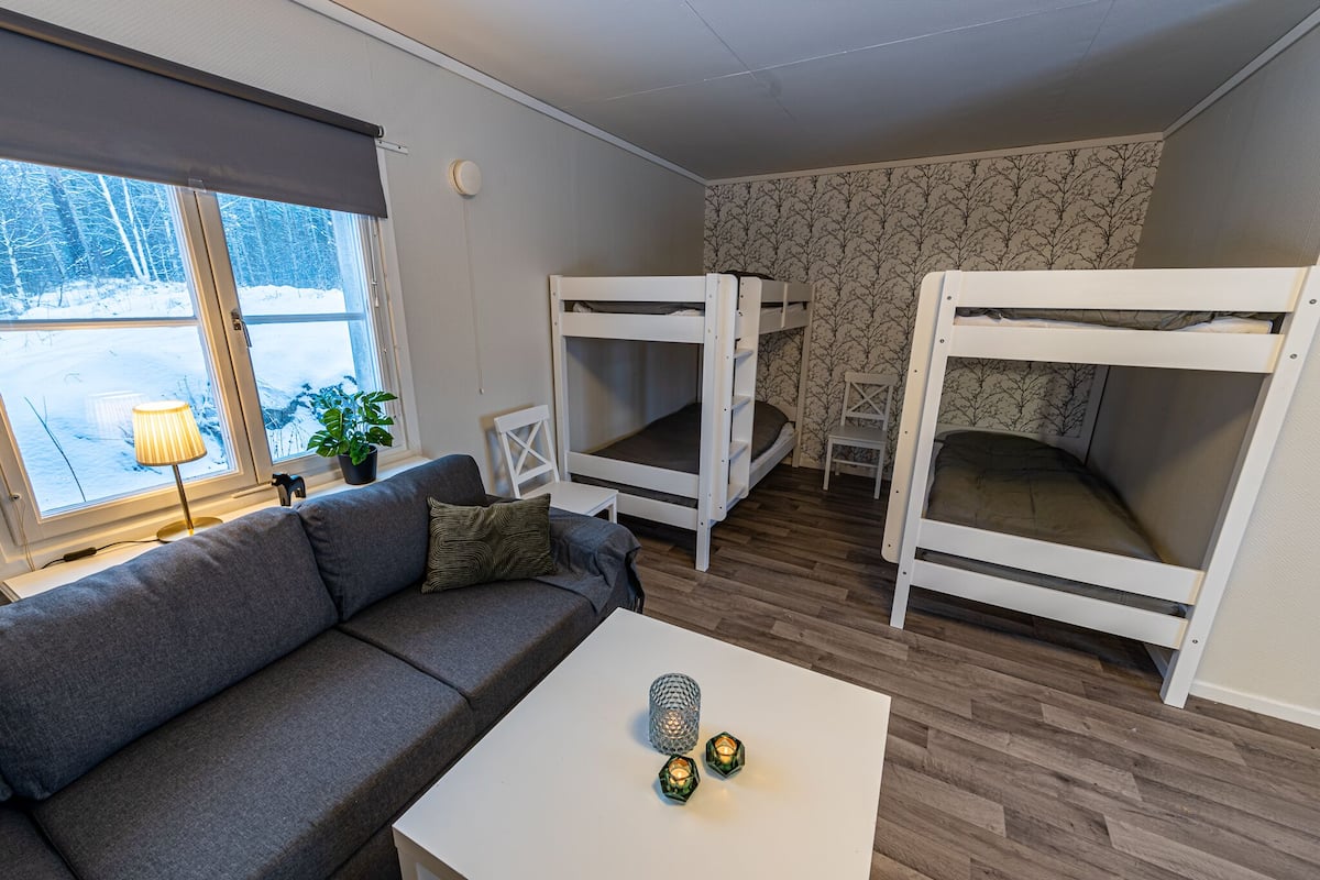 A cozy room with a grey sofa, two bunk beds, a white coffee table with candles, a lamp, and a window with a view of snow-covered trees.
