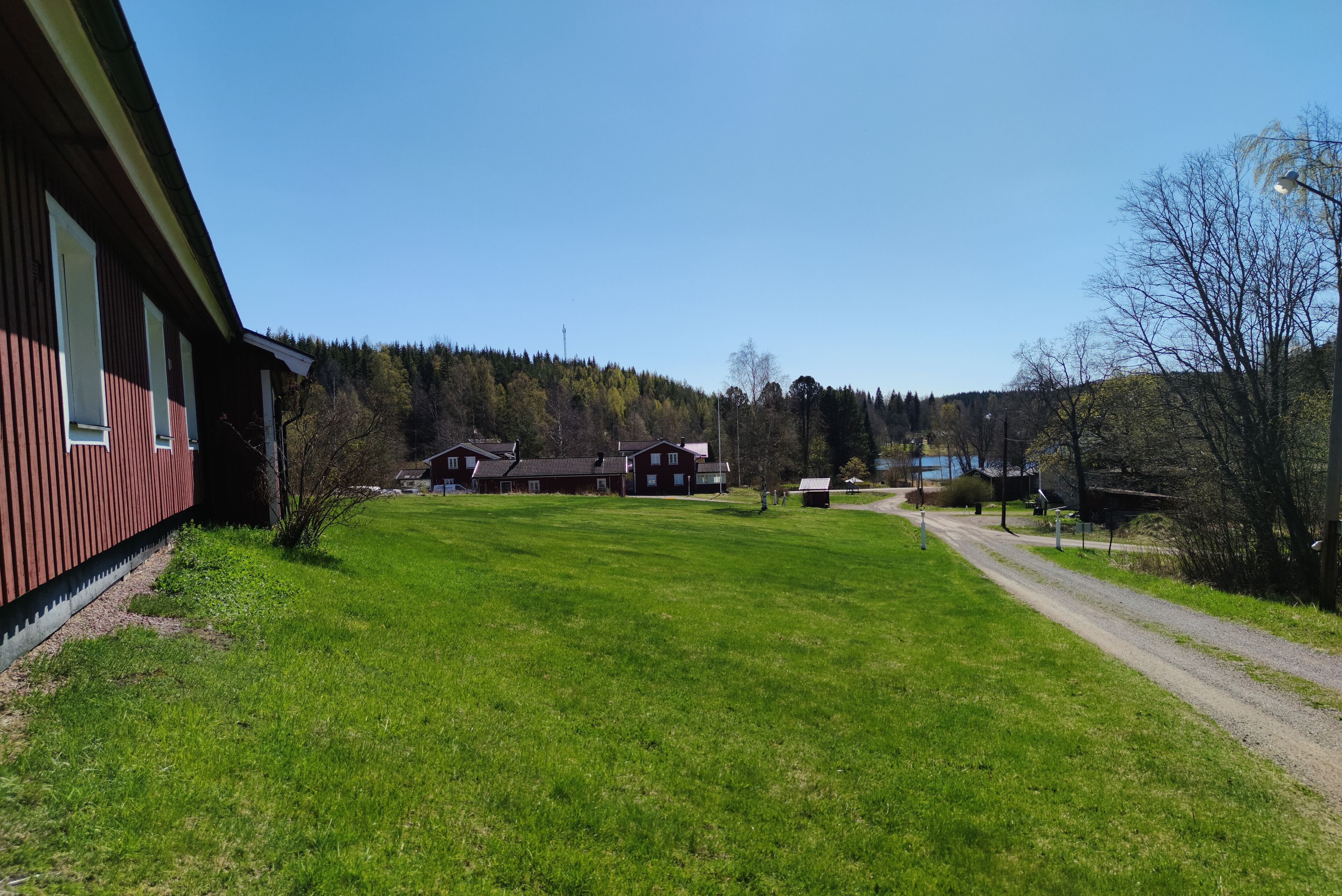 A building with a red exterior, a grassy yard, and a dirt path leading to a cluster of houses in the background.