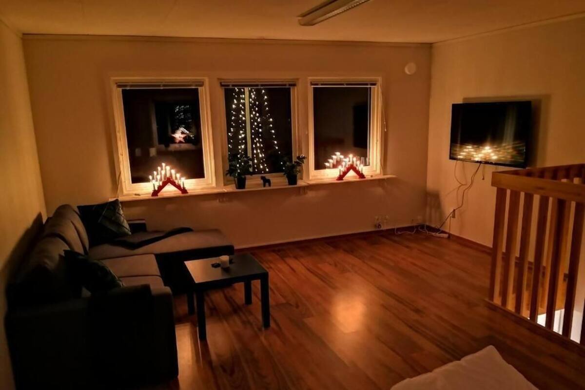 A living room with a sofa, a coffee table, and a TV mounted on the wall. There are three windows with Christmas lights and a small Christmas tree.