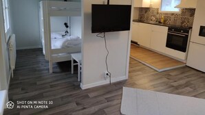 Habitación compacta con literas, un televisor de pantalla plana en la pared, una pequeña cocina con microondas y horno, y piso de madera.