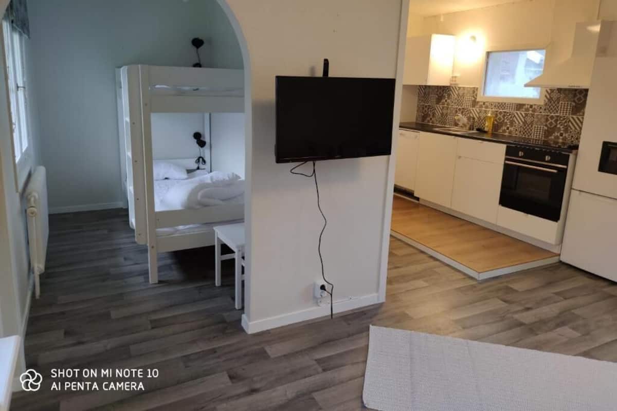 A compact room with a bunk bed, a flat-screen TV mounted on the wall, a small kitchen area with a microwave and oven, and a wooden floor.