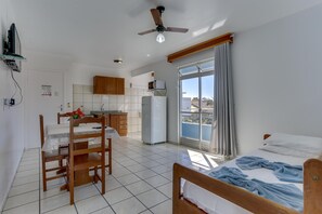 A compact bedroom with a bed, a small table, and a chair. Adjacent is a kitchenette with a refrigerator and a microwave. A ceiling fan is installed above the bed. - Marazul Apart Hotel II (Florianopolis)