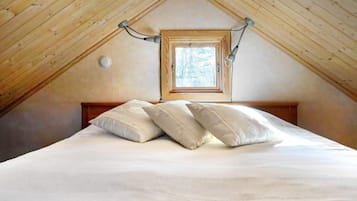 Ett sovrum med ett sluttande tak, ett fönster, en säng med kuddar och en väggmonterad lampa.