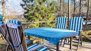 Een houten terras met een tafel en gestreepte stoelen, omringd door bomen en een gebouw op de achtergrond.