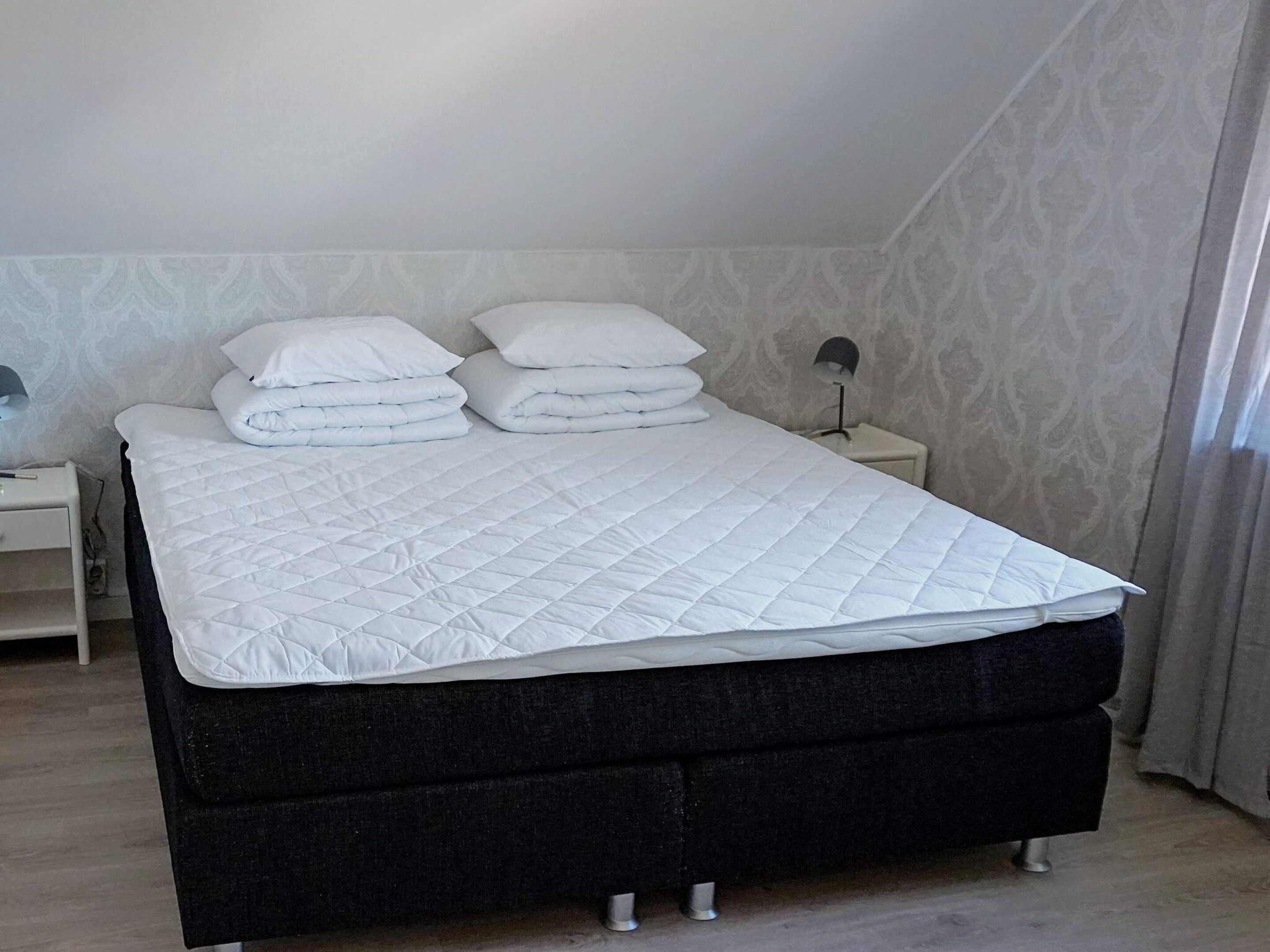 Siististi pedattu sänky, jossa on valkoinen peitto ja tyynyt, tumma pääty ja yöpöytä, jossa on lamppu.