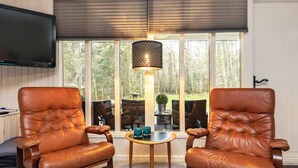 A room with two leather armchairs, a small wooden table, a TV mounted on the wall, and a large window with a view of trees. - 4 Person Holiday Home in Hals (Hals)