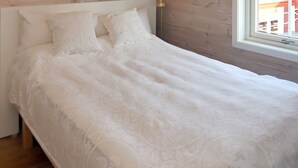 Un lit avec une couverture tricotée blanche et des oreillers, une tête de lit en bois et une fenêtre donnant sur des immeubles.
