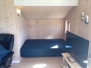 A small room with wooden walls, a single bed, a black chair, a TV, and a small table with a remote control.