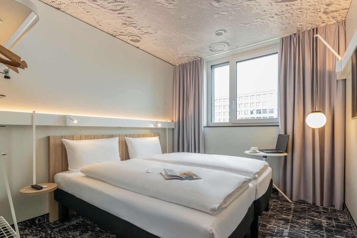 Une chambre d’hôtel avec un grand lit, un bureau, un ordinateur portable et une fenêtre avec des rideaux.
