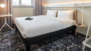 Une chambre d’hôtel avec un grand lit, une petite table de chevet, une lampe et une vue sur la ville par la fenêtre.