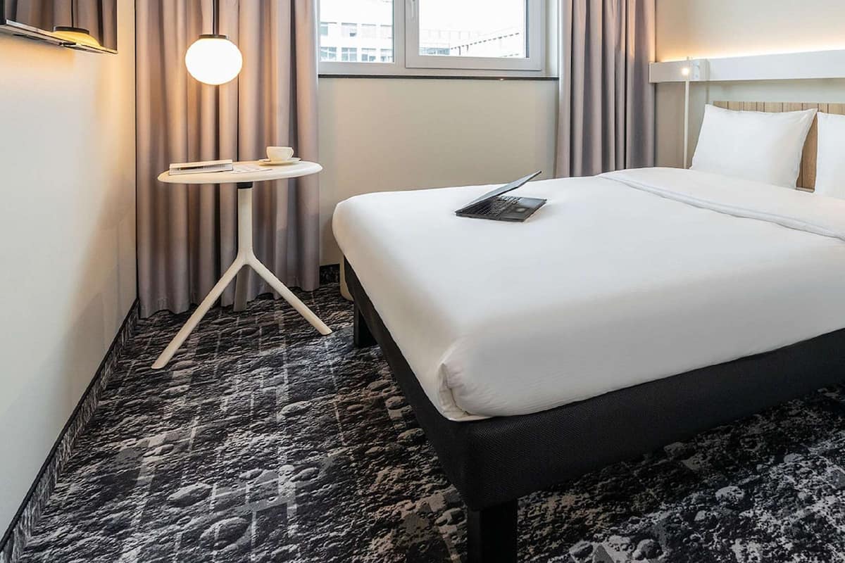 Une chambre d’hôtel avec un grand lit, une petite table de chevet, une lampe et une vue sur la ville par la fenêtre.