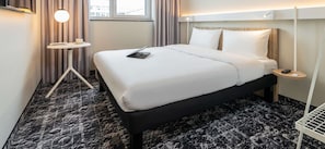 A hotel room with a large bed, a small side table, a lamp, and a view of the city through the window.