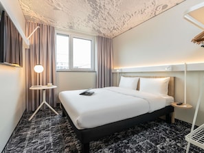 A hotel room with a bed, a desk, a chair, a window, and a ceiling with a unique pattern.
