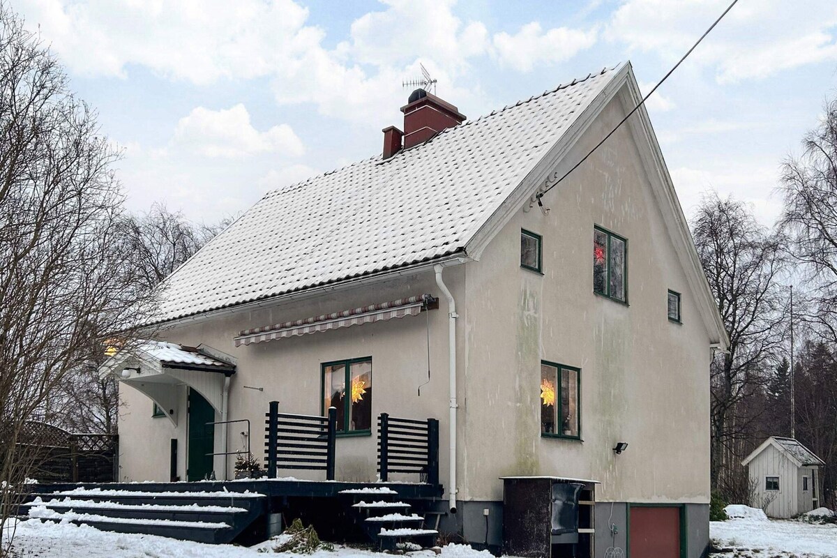 Uma casa de dois andares com telhado inclinado, varanda frontal com degraus e um pequeno galpão ao fundo.