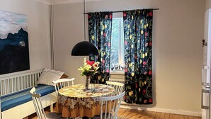 A cozy room with a round table, white chairs, a sofa with a blue cushion, a painting on the wall, and a window with floral curtains. - 7 Person Holiday Home in Docksta (Docksta)