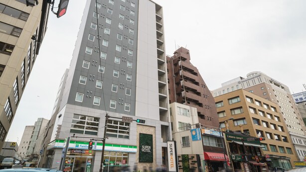 A multi-story hotel with a prominent 'Super Hotel' sign, a variety of shops on the ground floor, and a busy street with vehicles and pedestrians. - Super Hotel Yokohama Kannai (Yokohama)