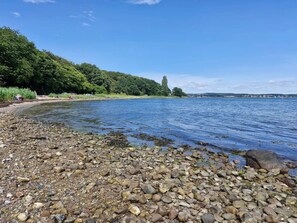 A pebbly shoreline with a calm body of water, surrounded by dense greenery and a clear sky. - 4 Star Holiday Home in Faaborg (Faaborg)