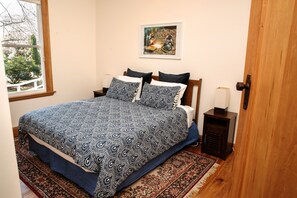 A bedroom with a large bed, a nightstand, a bedside table, a framed picture on the wall, and a window with a view of trees. - Elegant Villa Nestled In The Trees (Nelson)
