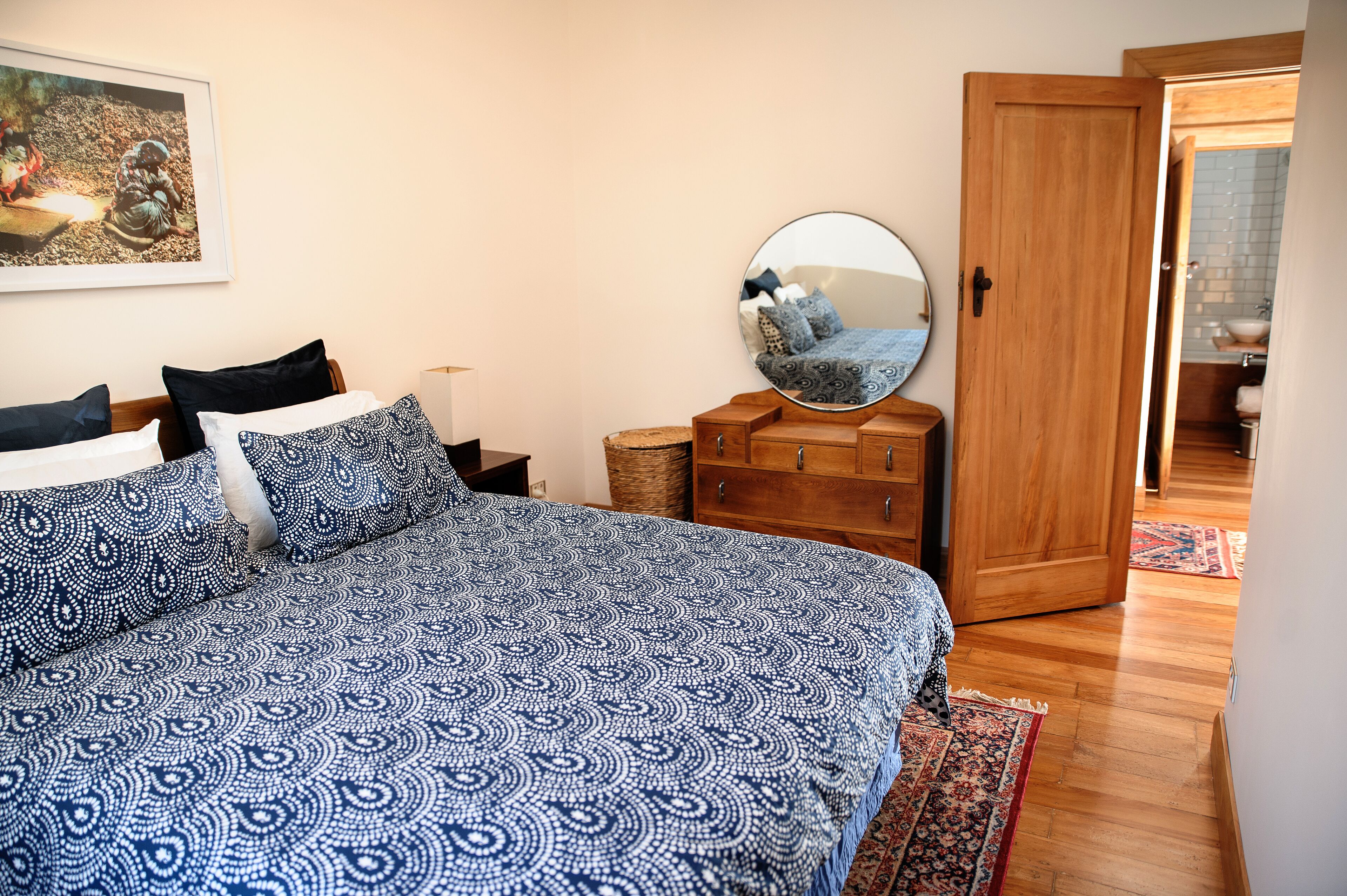 Een slaapkamer met een bed met een patroon, een houten dressoir, een ronde spiegel en een schilderij aan de muur.