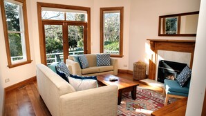 A living room with a sofa, armchair, and a fireplace. There are large windows and a view of trees outside. - Elegant Villa Nestled In The Trees (Nelson)