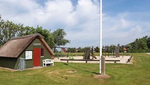 Ein kleines, grünes Gebäude mit roter Tür und einem Strohdach, gelegen in einem Park mit Spielgeräten und einem hohen, weißen Pfahl.