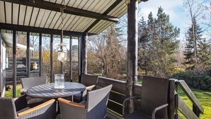 Un espacio al aire libre techado con mobiliario de mimbre y una mesa, con vista a un jardín con árboles y una terraza de madera.