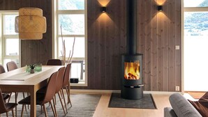A modern interior with a wood-paneled wall, a central wood-burning stove, a dining area with chairs and a table, and large windows offering a view of the outdoors. - 8 Person Holiday Home in Skei i Jolster (Sunnfjord)