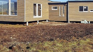 Um edifício moderno, térreo, com amplas janelas e telhado plano, localizado em terreno desimpedido com vegetação rala.